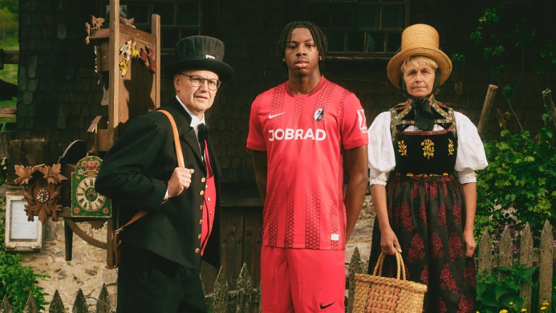 Ein SC Spieler steht vor dem Hof, daneben 2 historische Kaufleute mit Kuckucksuhren Ein SC Spieler steht vor dem Hof, daneben 2 historische Kaufleute mit Kuckucksuhren